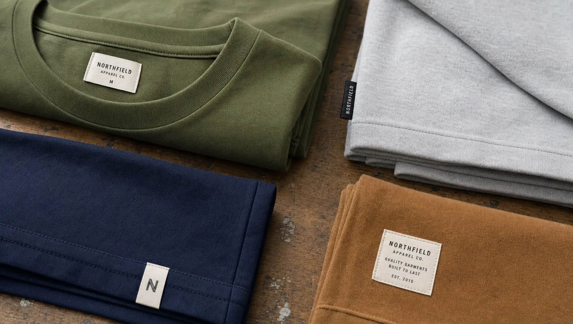 A close-up of several folded apparel pieces on a worktable, each revealing a different woven tag label placement such as a neck label, side seam tag, hem tag, and woven patch.