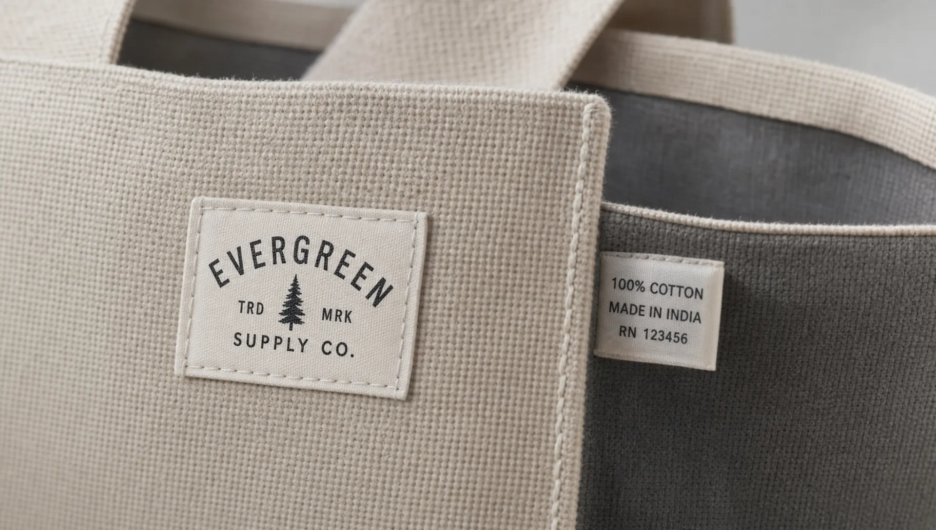 A close-up view of a woven label patch sewn onto the outside front corner of a textured canvas tote bag, with a small standard woven tag peeking from the inner seam to show the contrast between visible branding and interior identification.