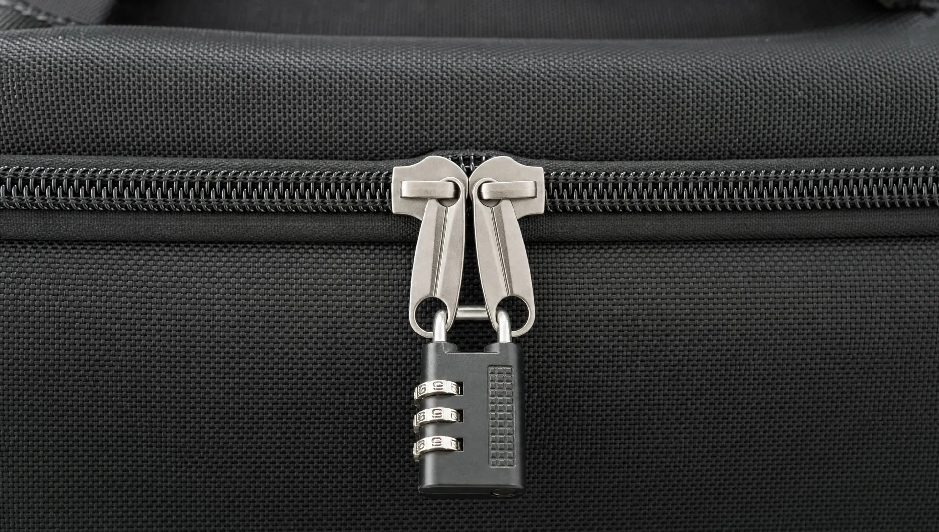 A close-up of interlocking zipper pulls on a travel bag, with two aligned tabs meeting at the center of a closed zipper and a small travel lock passing through both openings.
