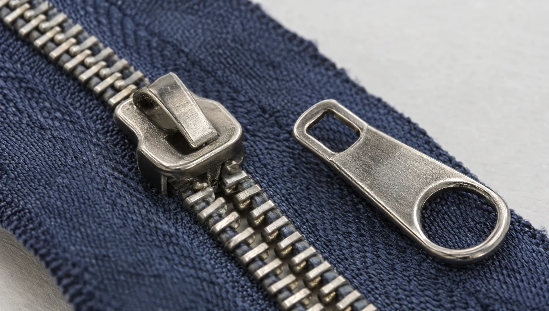 A zipper slider fitted around a zipper chain sits beside a separate pull tab, with the connection point visible against the fabric.