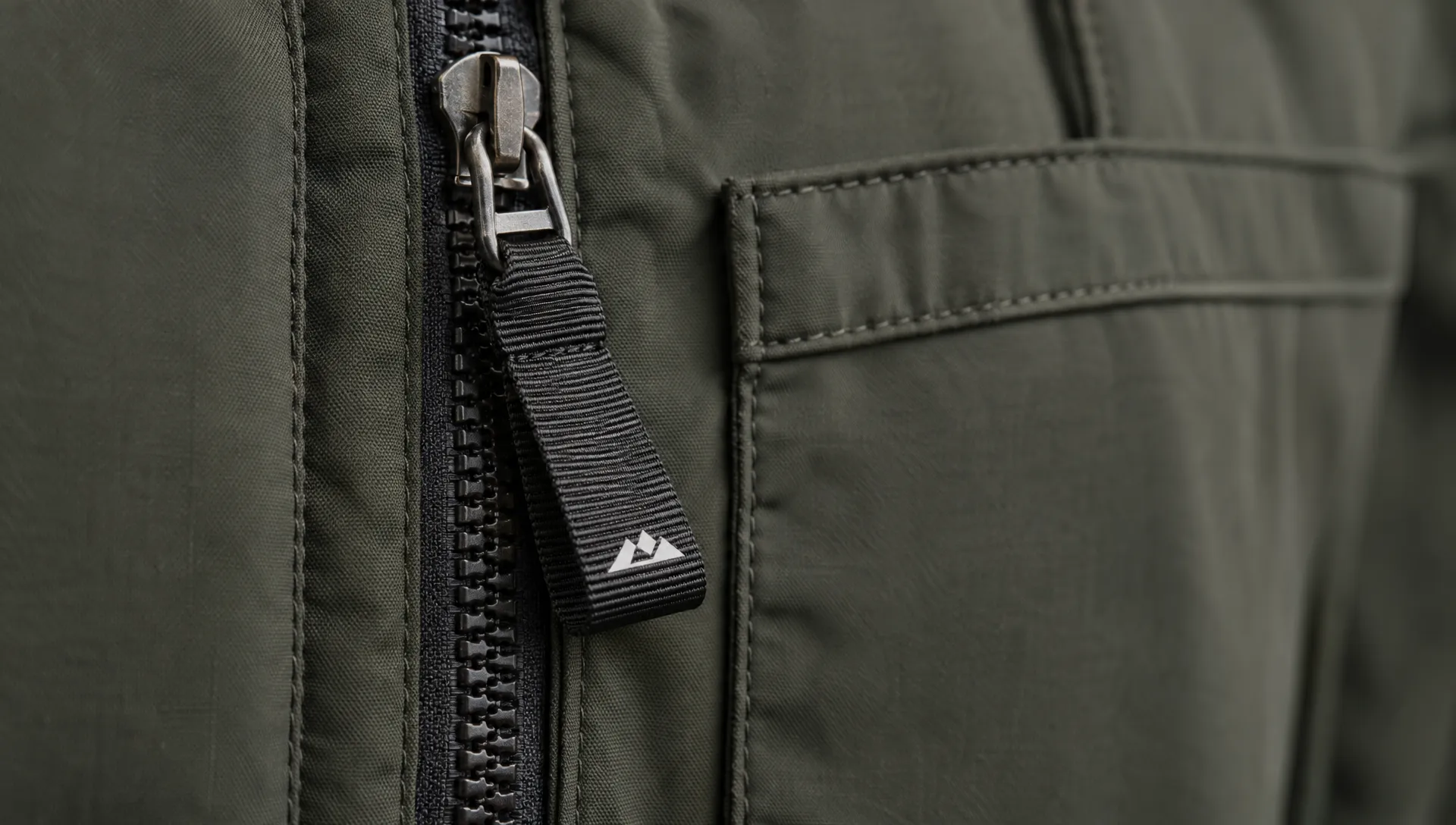 Grosgrain ribbon with a compact brand mark forms a folded pull on a fabric jacket pocket, clear of the metal zipper slider.