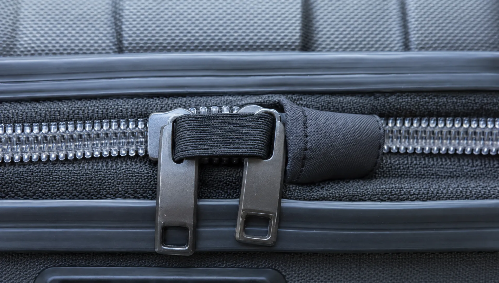 Luggage zipper pulls sit side by side under a removable elastic loop, while a fabric cuff cushions one metal tab beside the clear zipper teeth.