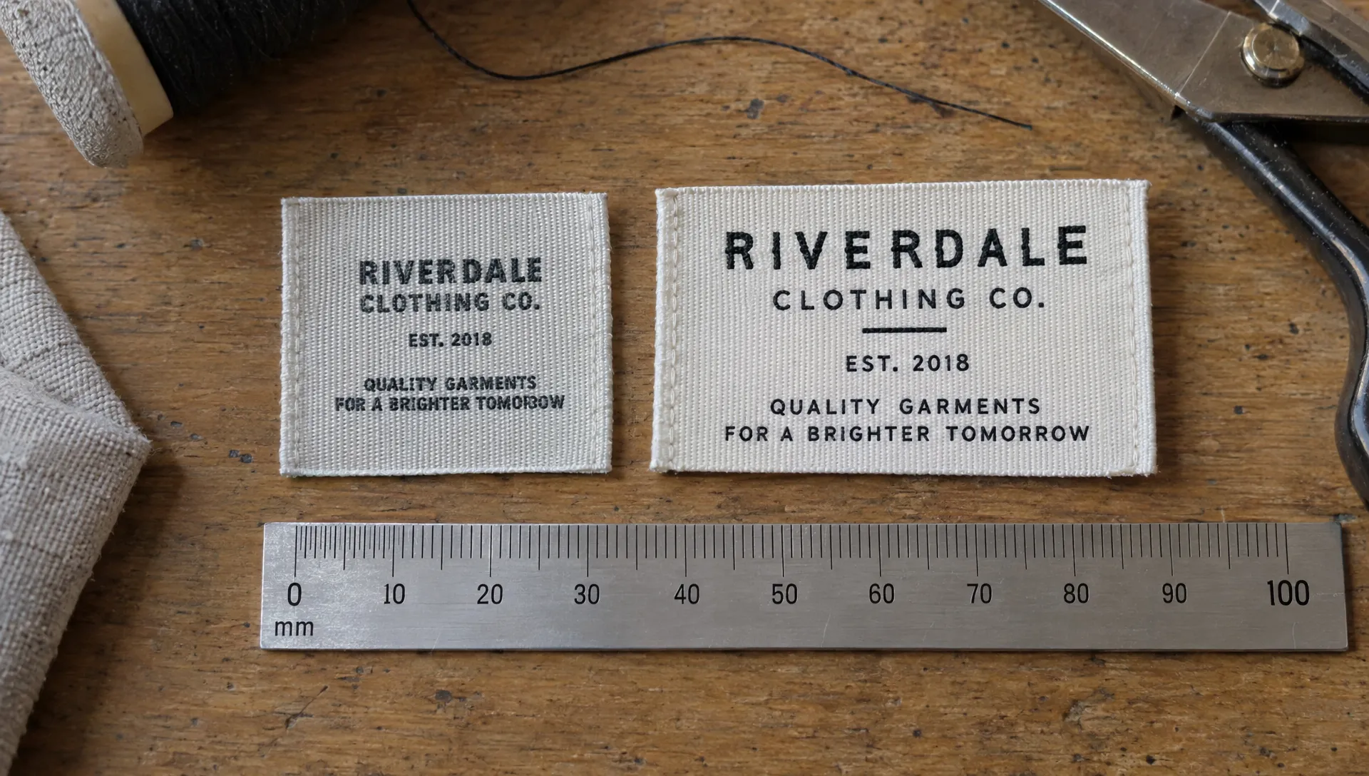 Two woven labels beside a millimeter ruler compare crowded tiny lettering with larger, more open lettering for woven label design.